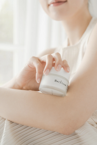 Woman holding jar of Herbloom Kombucha Vegan Collagen Cream on her arm, with only the bottom half of her face visible. She is wearing white and the curtains behind her are white.