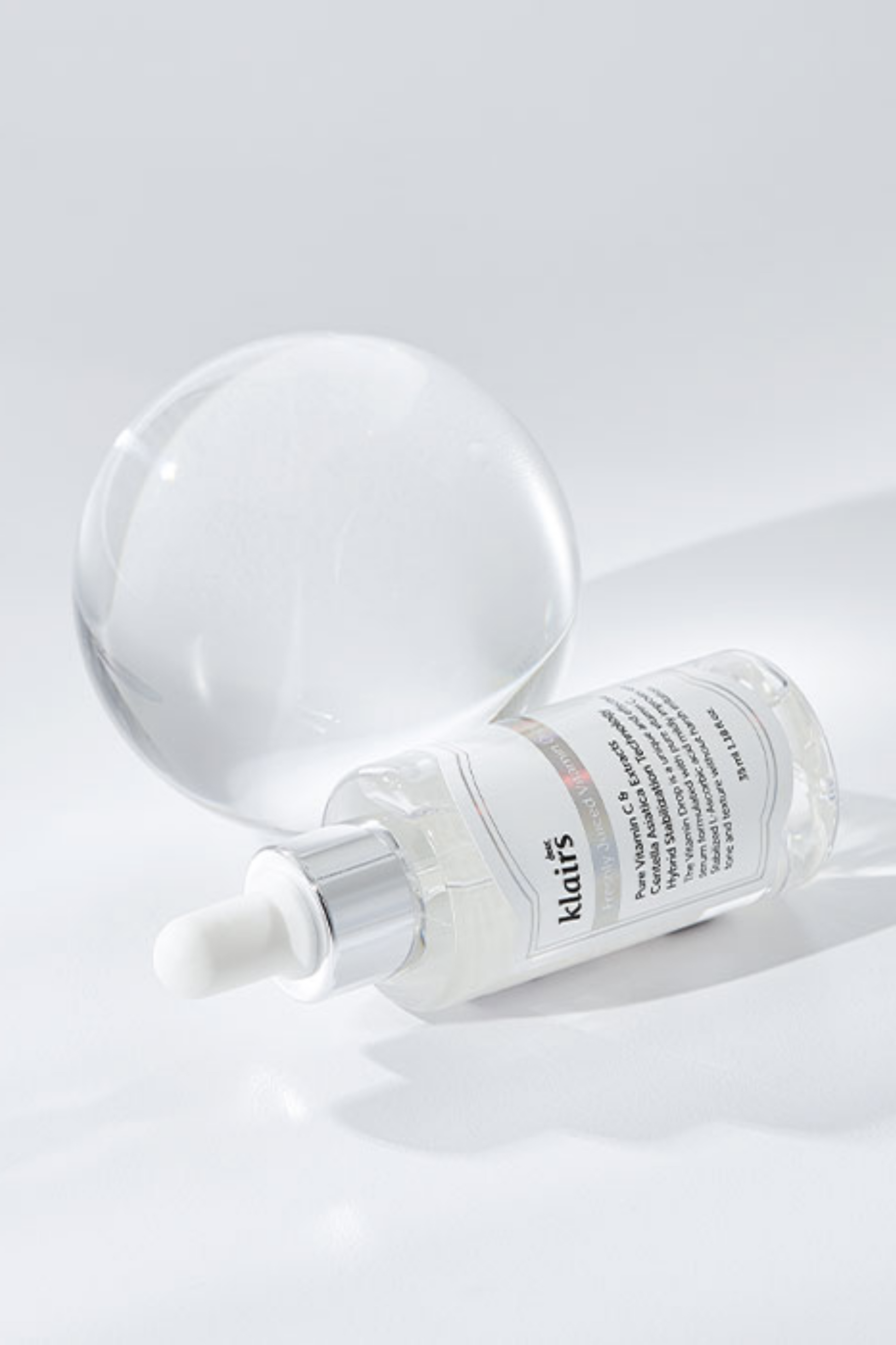 Bottle of Klairs' Freshly Juiced Vitamin Drop lying on its side in front of a glass sphere, against a light grey background.