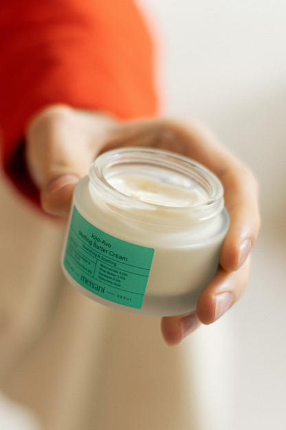 An open jar of Meisani's Jojo-Avo Melting Butter Cream in someone's hand with a blurred background. 