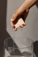 Load image into Gallery viewer, Woman&#39;s wet hand holding Ondo Beauty 36.5&#39;s Calamind &amp; Oatmeal Soothing Cleansing Bar with water dripping down, against a grey background.
