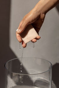 Woman's wet hand holding Ondo Beauty 36.5's Calamind & Oatmeal Soothing Cleansing Bar with water dripping down, against a grey background.