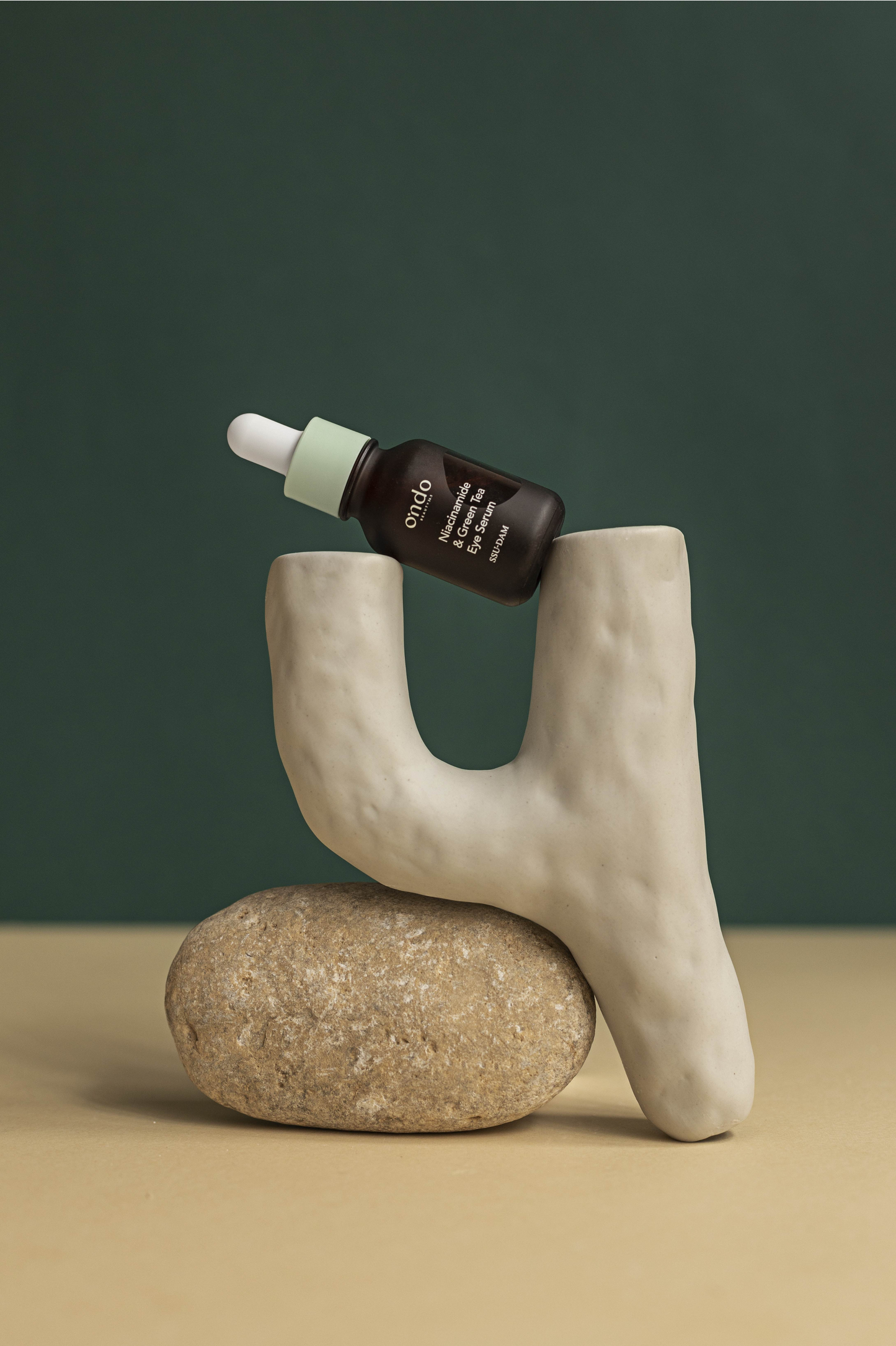 Ondo Beauty 36.5 Green Tea Eye Serum laying sideways on a white stone against a dark green background. Available for purchase at Sinless Skincare.