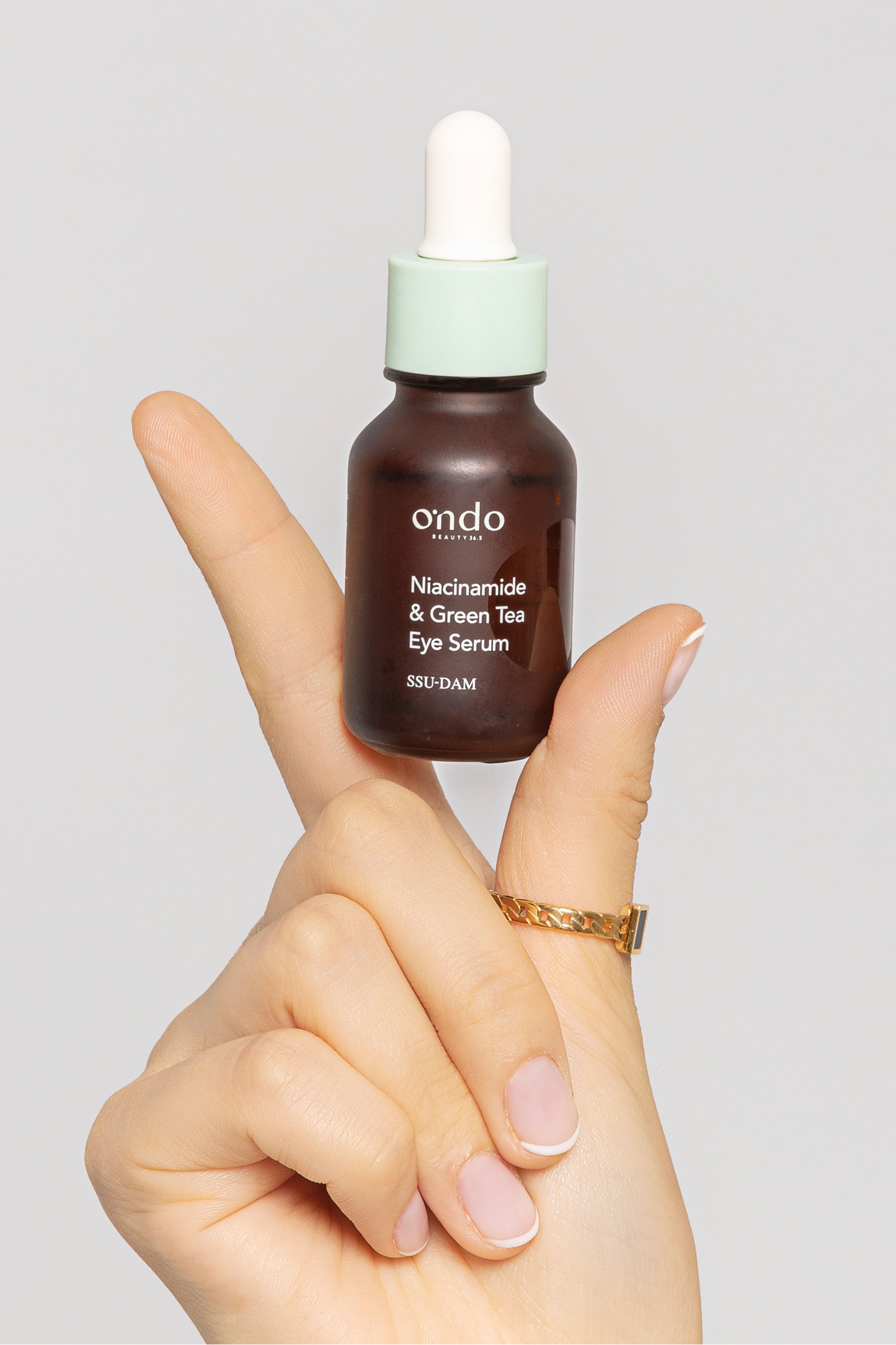 Close up of a woman's hand holding up a bottle of Ondo Beauty 36.5 Green Tea Eye Serum, against a light grey background. Available for purchase at Sinless Skincare.