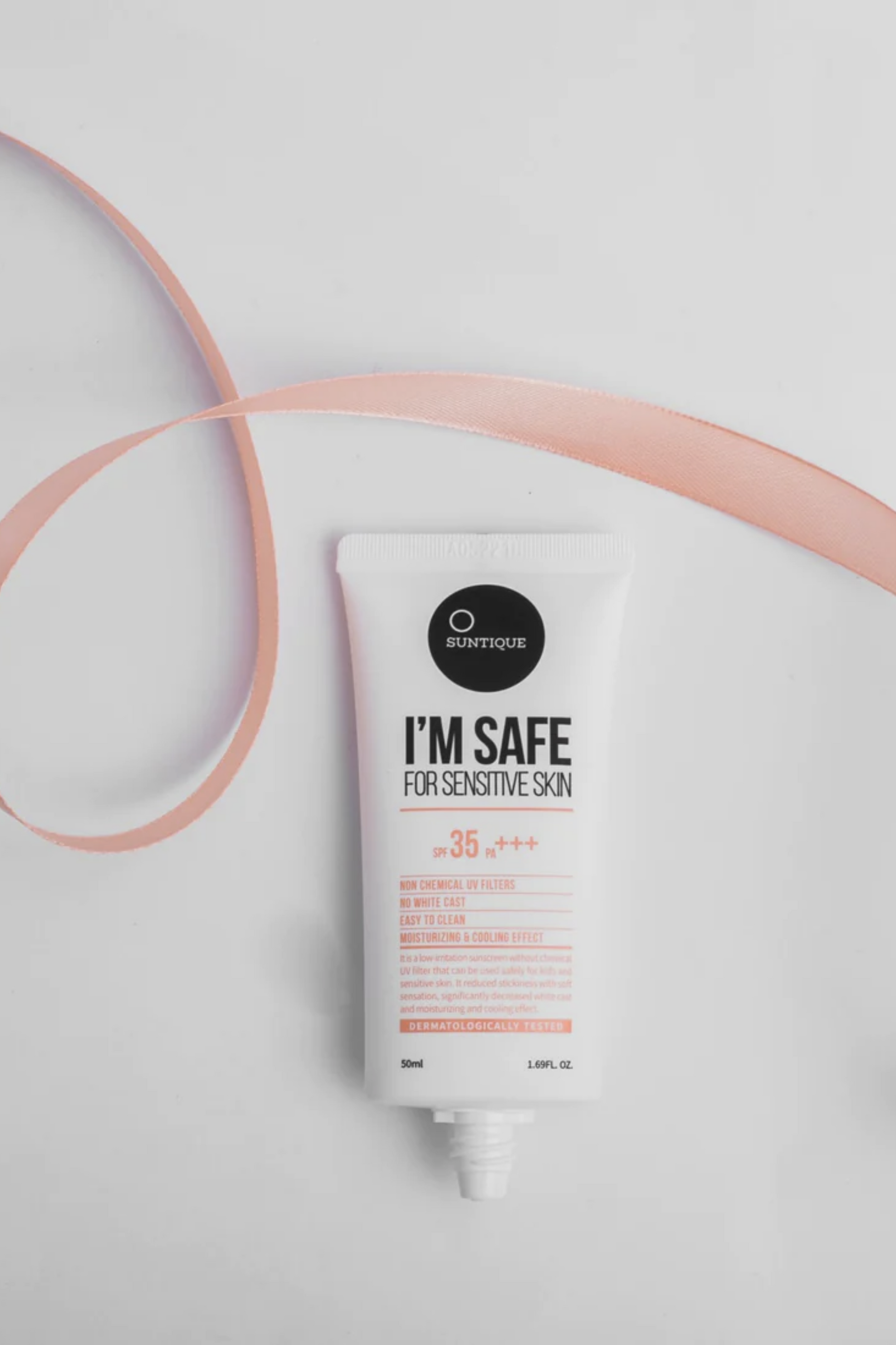 Tube of Suntique's I'm Safe for Sensitive Skin without its cap. A light pink ribbon is laying above the tube. Against a light grey background. Available for purchase at Sinless Skincare.