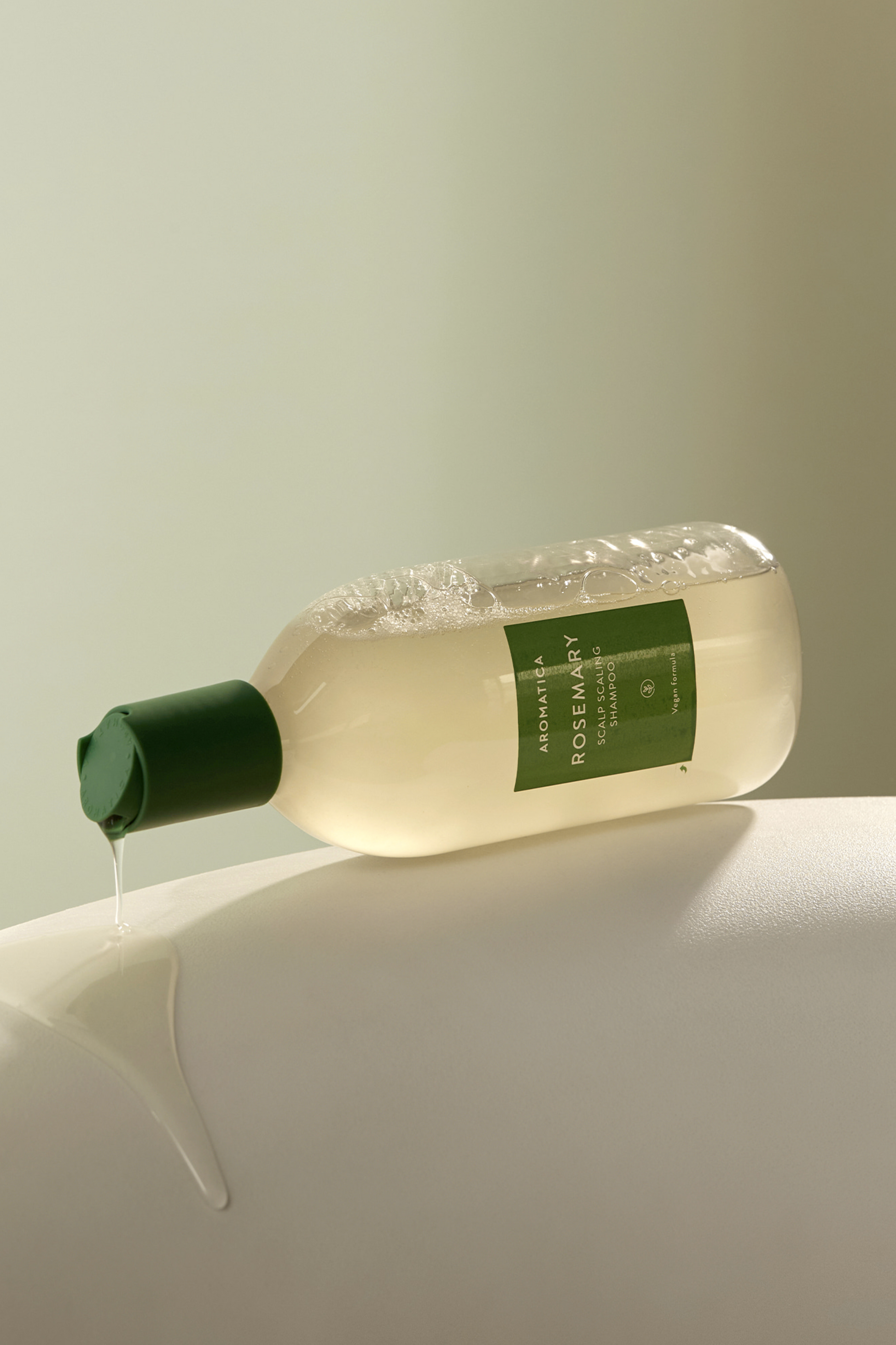 400ml bottle of Aromatica's Rosemary Scalp Scaling Shampoo tipped on its side with shampoo dripping down from its cap. 