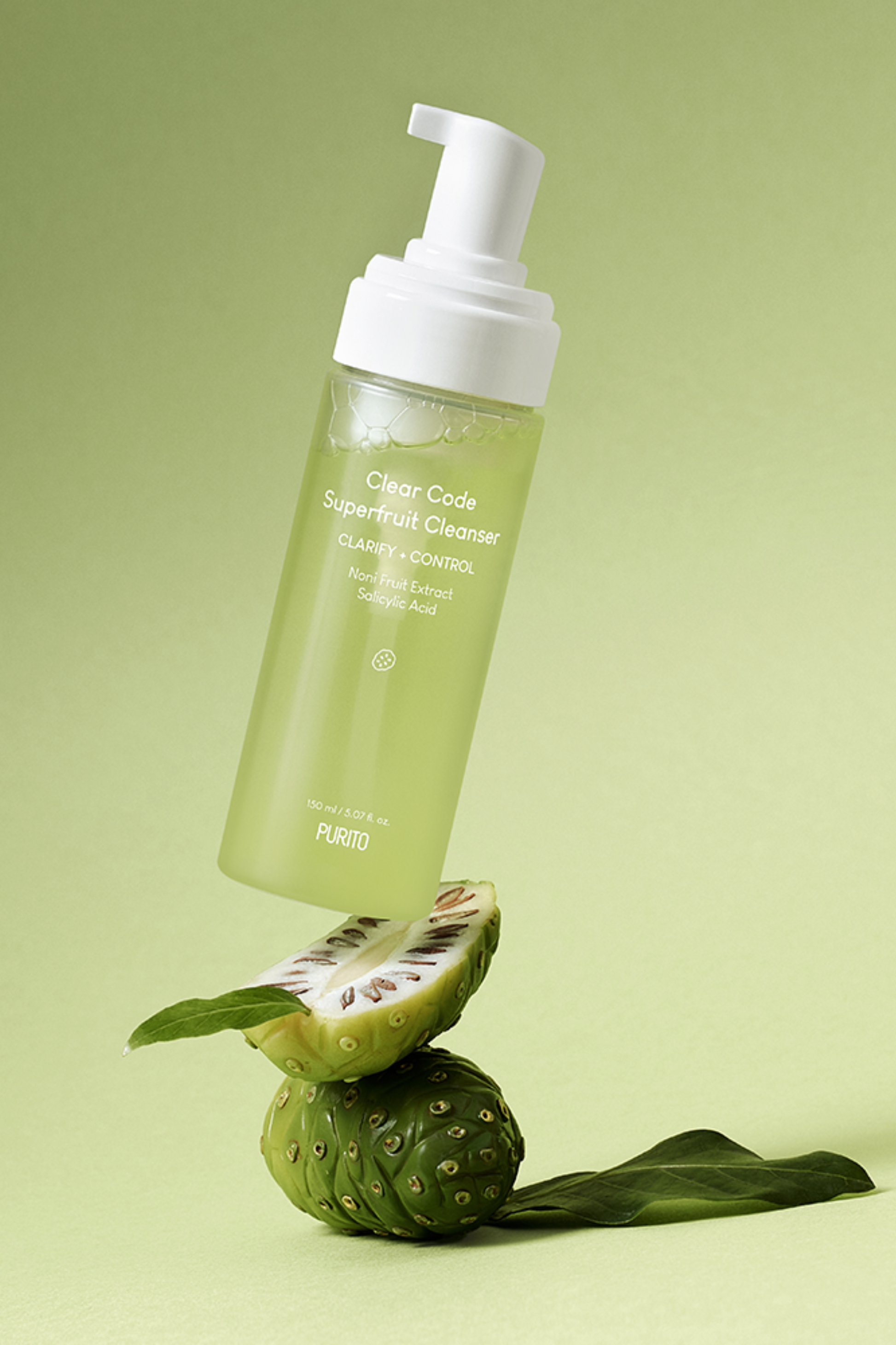 Capless bottle of Purito's Clear Code Superfruit Cleanser on top of two pieces of Noni fruit, against a light green background. Available for purchase at Sinless Skincare.