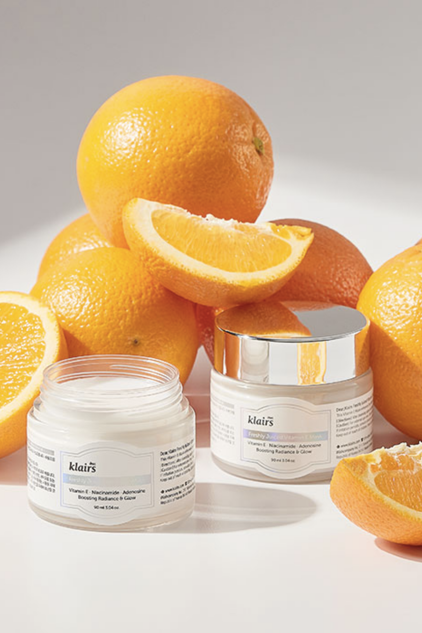 Two jars of Klairs' Freshly Juiced Vitamin E Mask in front of a pile of oranges, against a light grey background. One jar is without its lid and the cream is visible.