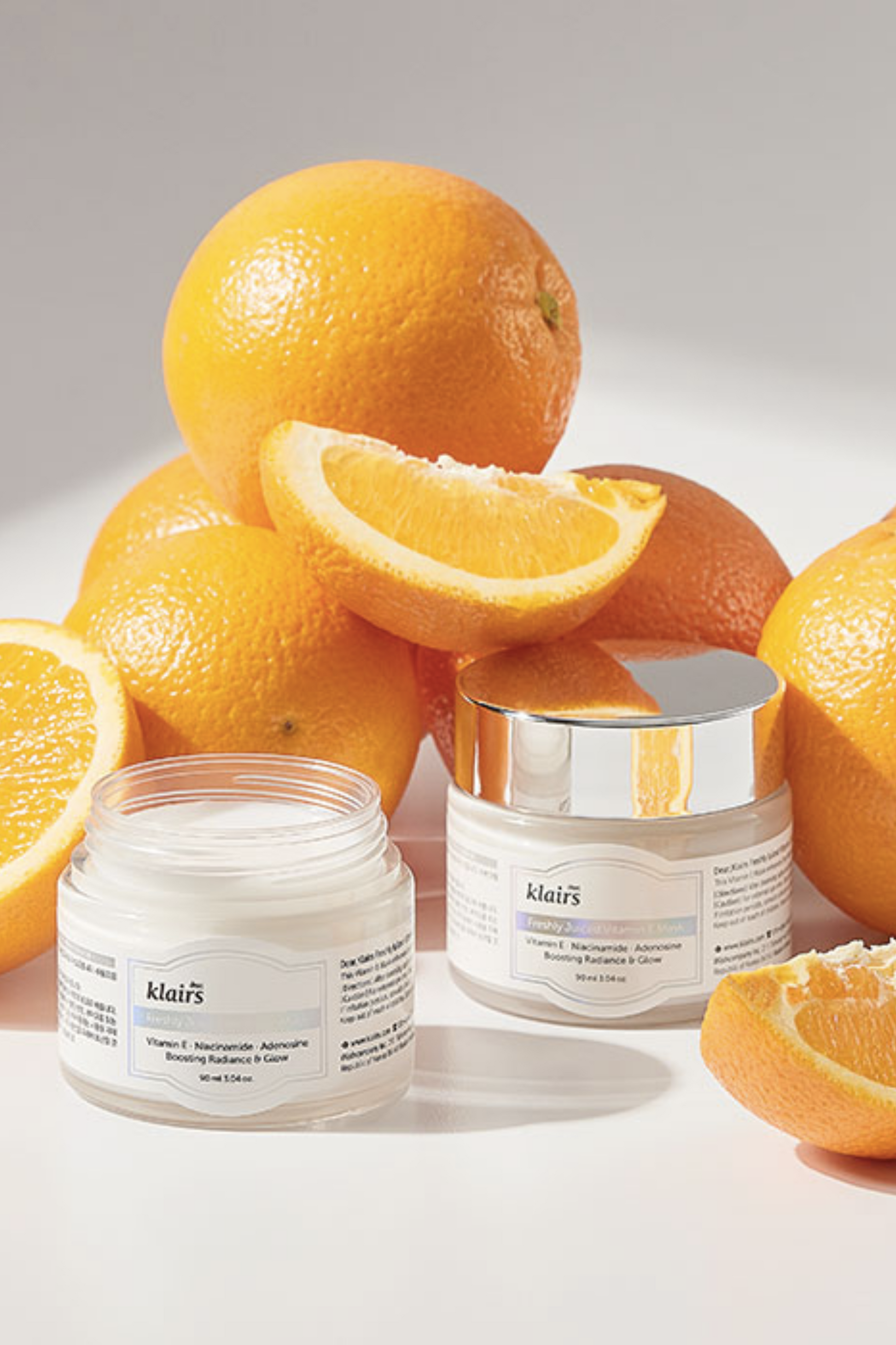 Two jars of Klairs' Freshly Juiced Vitamin E Mask in front of a pile of oranges, against a light grey background. One jar is without its lid and the cream is visible.