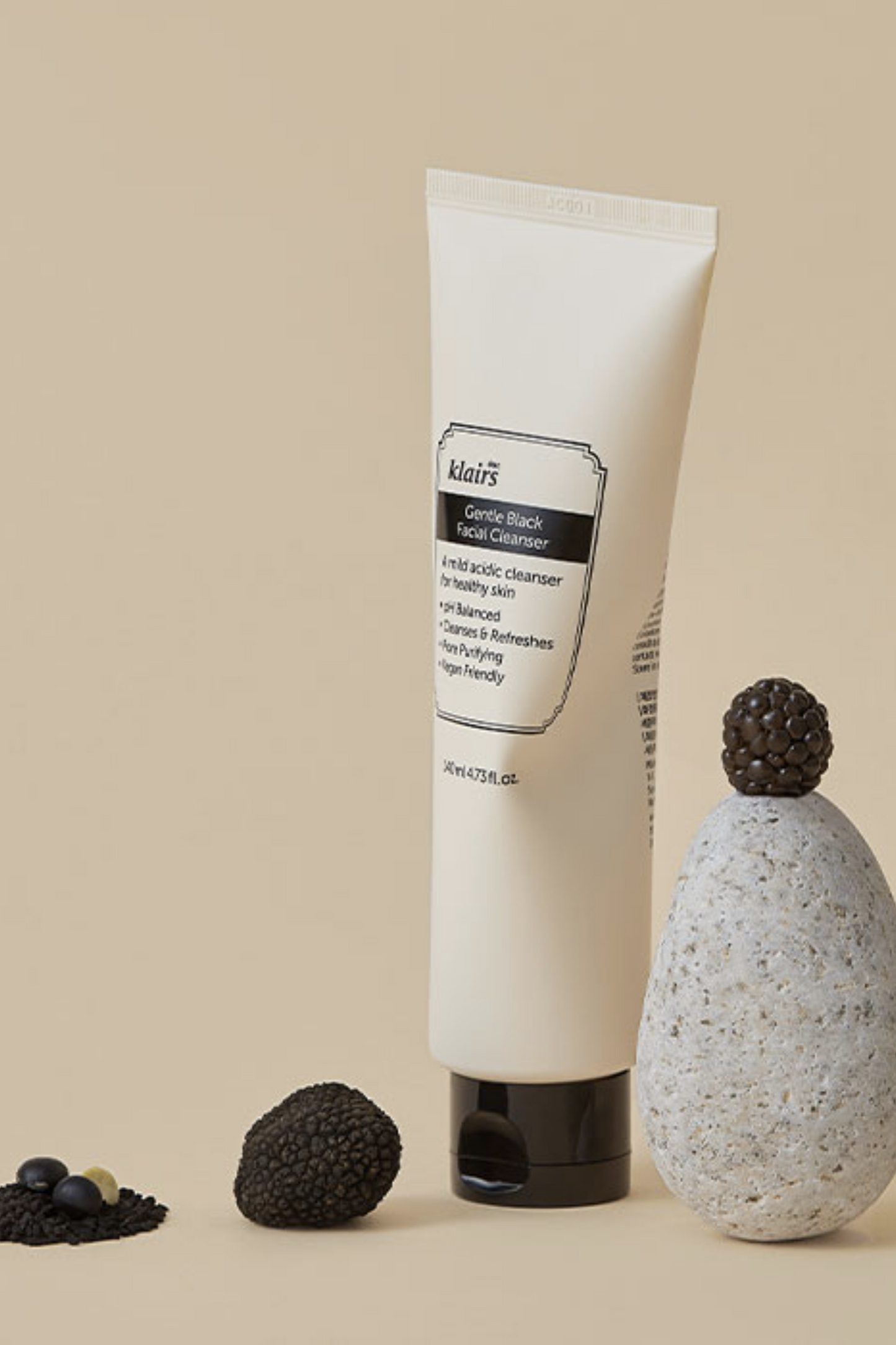 Upright, visibly squeezed tube of Klairs Gentle Black Facial Cleanser against a beige background. A grey stone with a blackberry on top is next to the tube. Blackberries are on the ground.