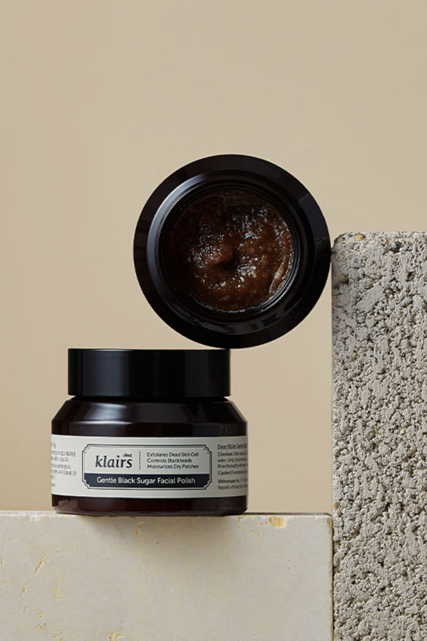 Two jars of Klairs Gentle Black Sugar Facial Polish. One is standing upright and the other is on top of it, on its side with its lid off so the exfoliator is visible. It is leaning against a grey stone with a beige background.