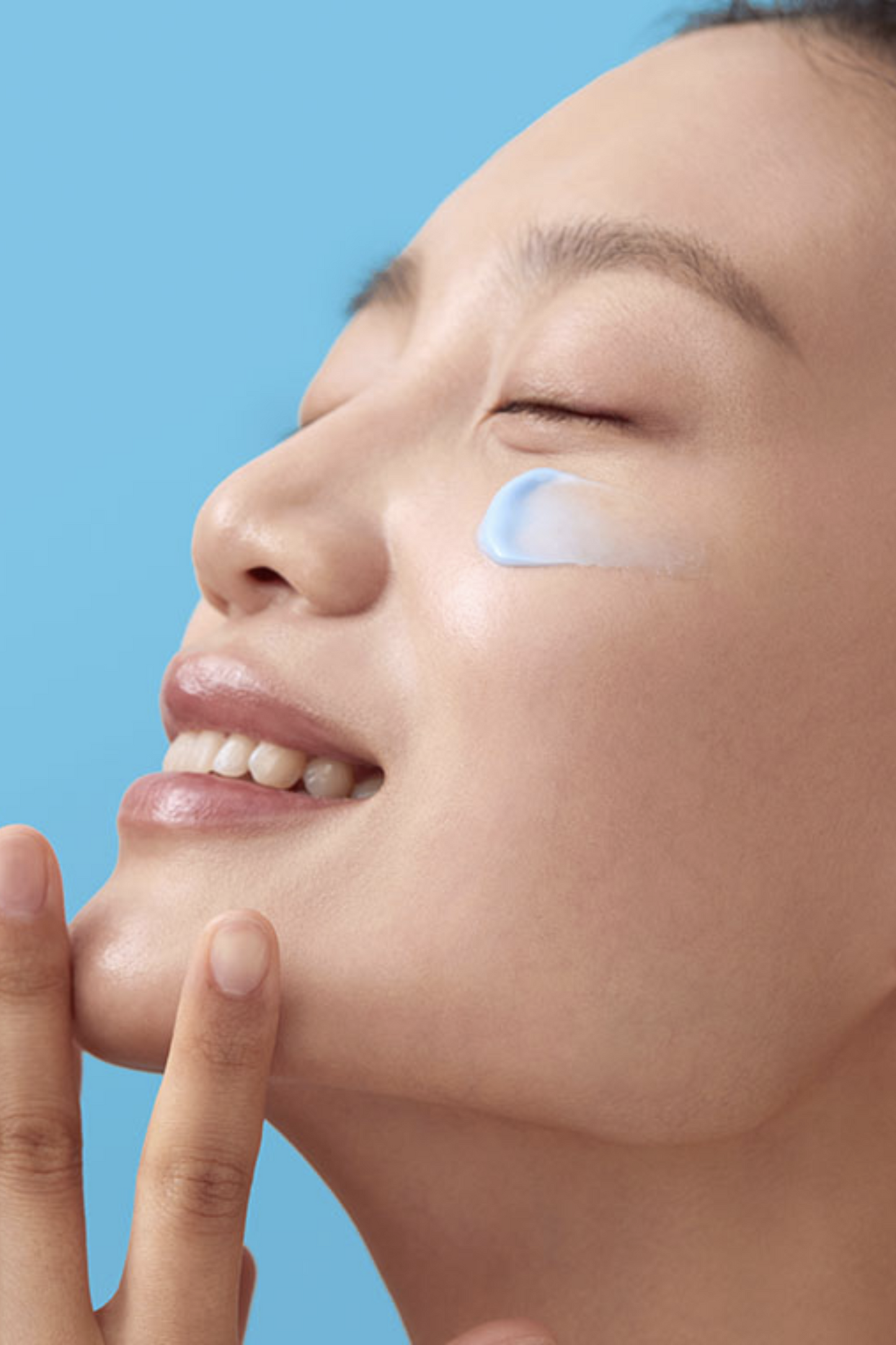 Side view of smiling woman's face and a smear of Klairs Midnight Blue Calming Cream below her eye, against a blue background.