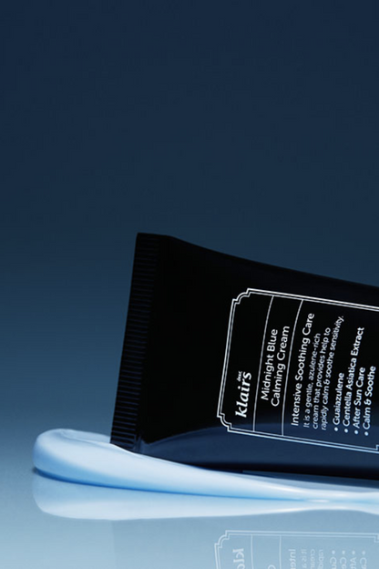 Tube of Klairs Midnight Blue Calming Cream laying on its side and on top of blue cream, against a dark blue background.