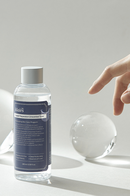 Bottle of Klairs' Supple Preparation Unscented Toner next to a glass ball, against a light grey background. Three fingers from a woman's hand are hovering over the ball.