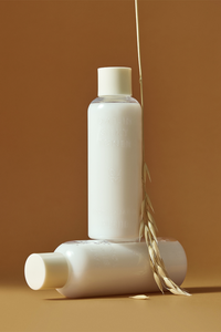 Two bottles of Purito's Oat-in Silky Toner. One is tipped sideways with the other standing on top of it. A piece of wheat is leaning against them, against a brown background.