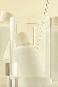 Close up of a bottle of Purito's Oat-in Silky Toner inside a glass, with other bottles of product standing behind it, against a beige background. Available for purchase at Sinless Skincare.