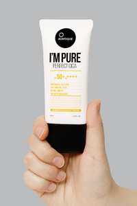 Close up of a hand holding up a tube of Suntique's I'm Pure Perfect Cica sunscreen against a light grey background. Available for purchase at Sinless Skincare.