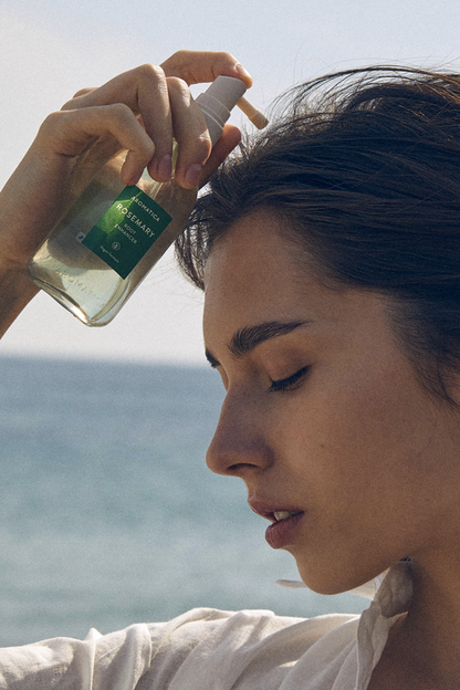 Profile view of woman standing at the sea, holding a bottle of Aromatica's Rosemary Root Enhancer to her scalp.