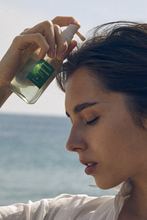 Load image into Gallery viewer, Profile view of woman standing at the sea, holding a bottle of Aromatica&#39;s Rosemary Root Enhancer to her scalp.
