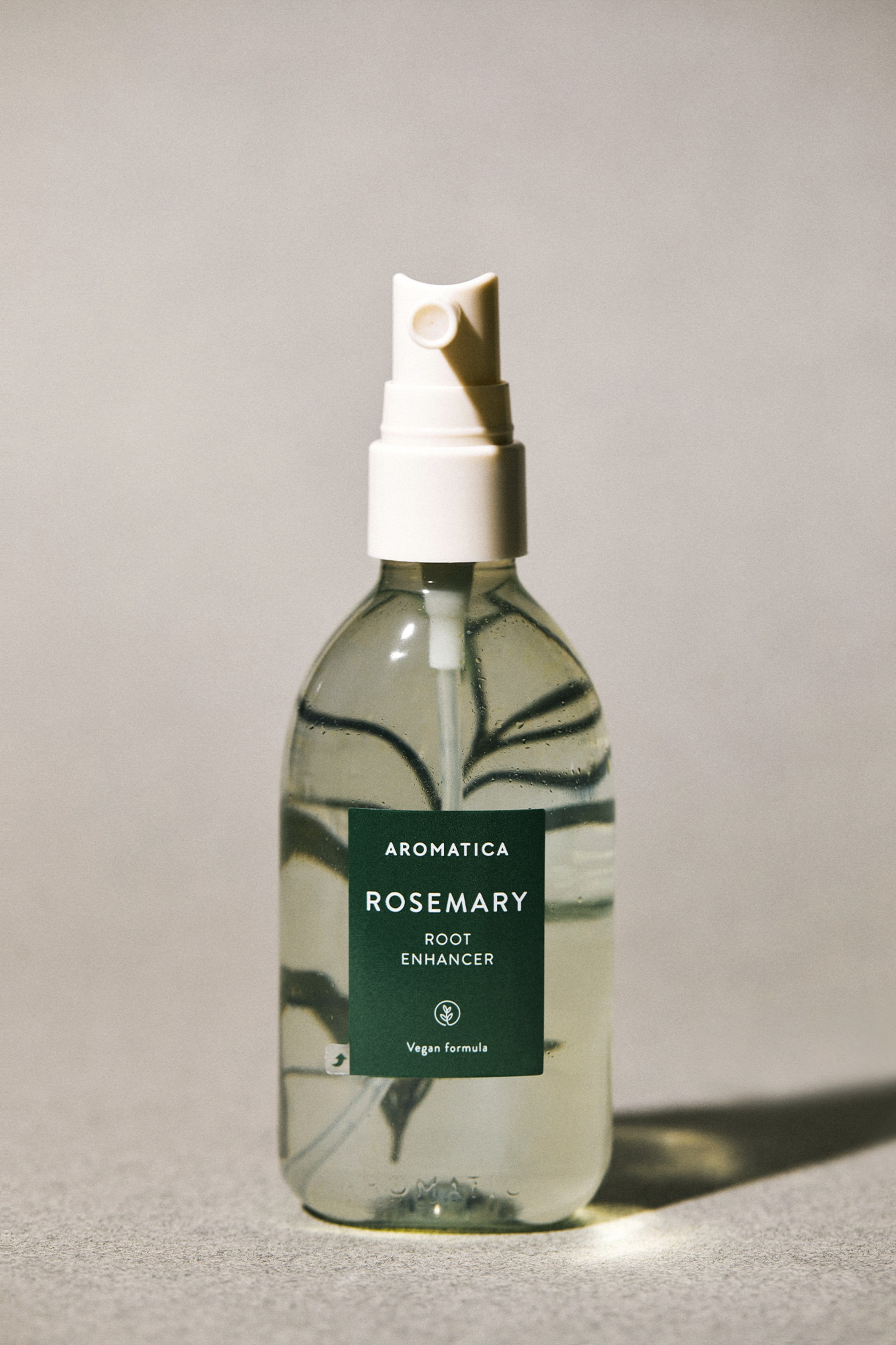 Front view of Aromatica's Rosemary Root Enhancer against a soft grey background, with a twig of Rosemary magnified from behind the transparent bottle.