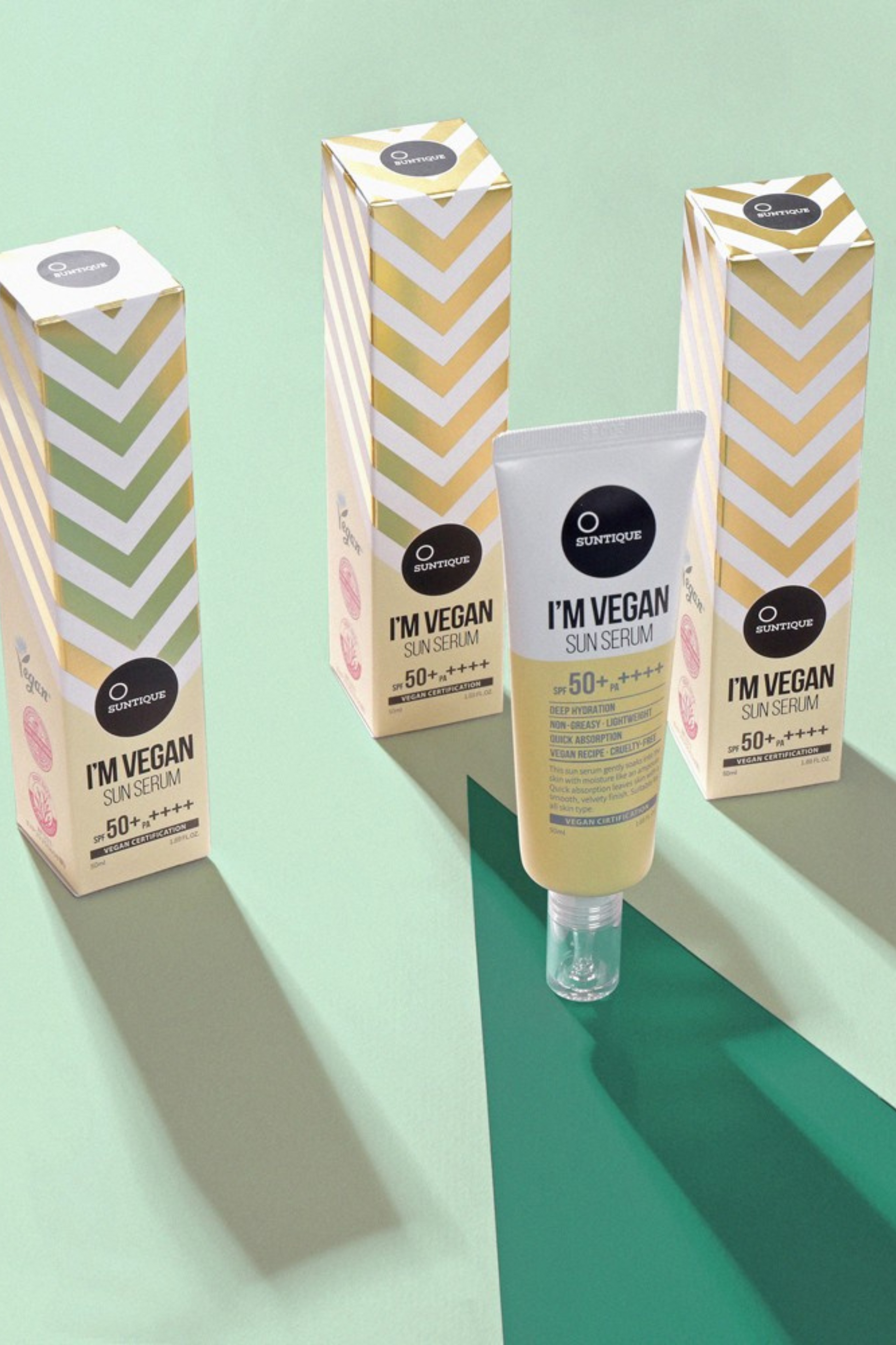 3 boxes of Suntique's I'm Vegan Sun Serum sunscreen standing up on light green background. One tube of product is standing in front outside of its box.