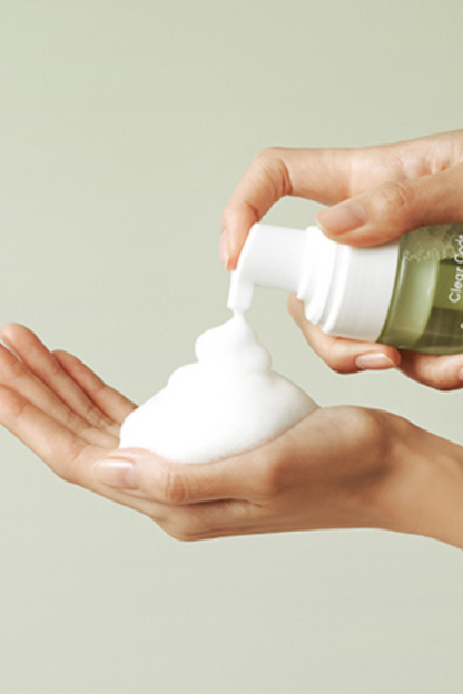 Close up of women's hands. She is pumping foam from a bottle of Purito's Clear Code Superfruit Cleanser onto her hand, against an off-white background. Available for purchase at Sinless Skincare.