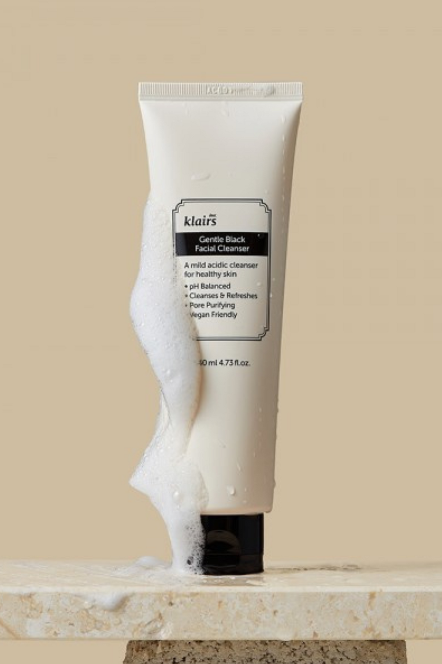 Upright bottle of Klairs Gentle Black Facial Cleanser with foam bubbles along its side. Bottle is standing on a stone platform against a beige background.