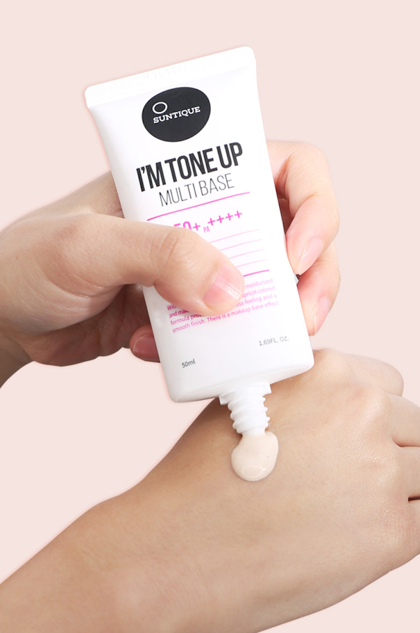 Tube of Suntique's Tone Up Multibase sunscreen being applied to someone's hand, against a beige background. Available for purchase at Sinless Skincare.